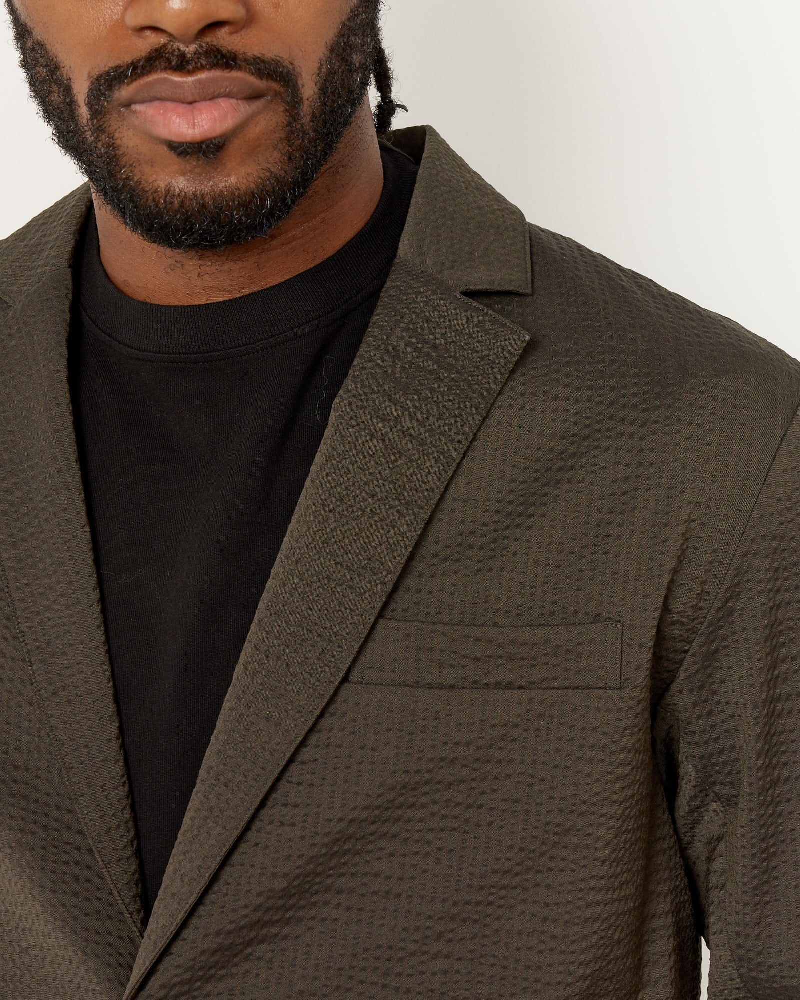 Close-up of a man wearing the SMOCK Blazer Super 120s Wool Seersucker in Olive over a black crew-neck shirt, showing his beard and upper torso against a plain light background.