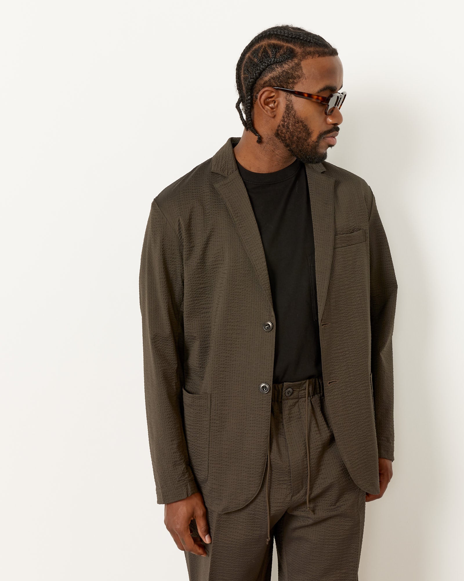 A man with braided hair and sunglasses wears the SMOCK Blazer Super 120s Wool Seersucker in Olive over a black t-shirt and matching trousers, standing against a plain white background.