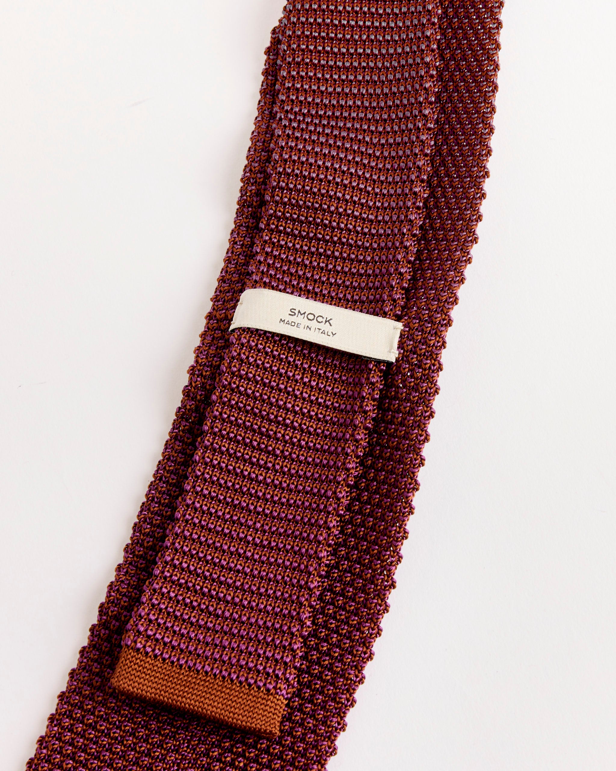 A close-up of the SMOCK Silk Knit Tie in Eggplant/Cognac, featuring a textured finish and a white SMOCK MADE IN ITALY label, displayed on a white background.