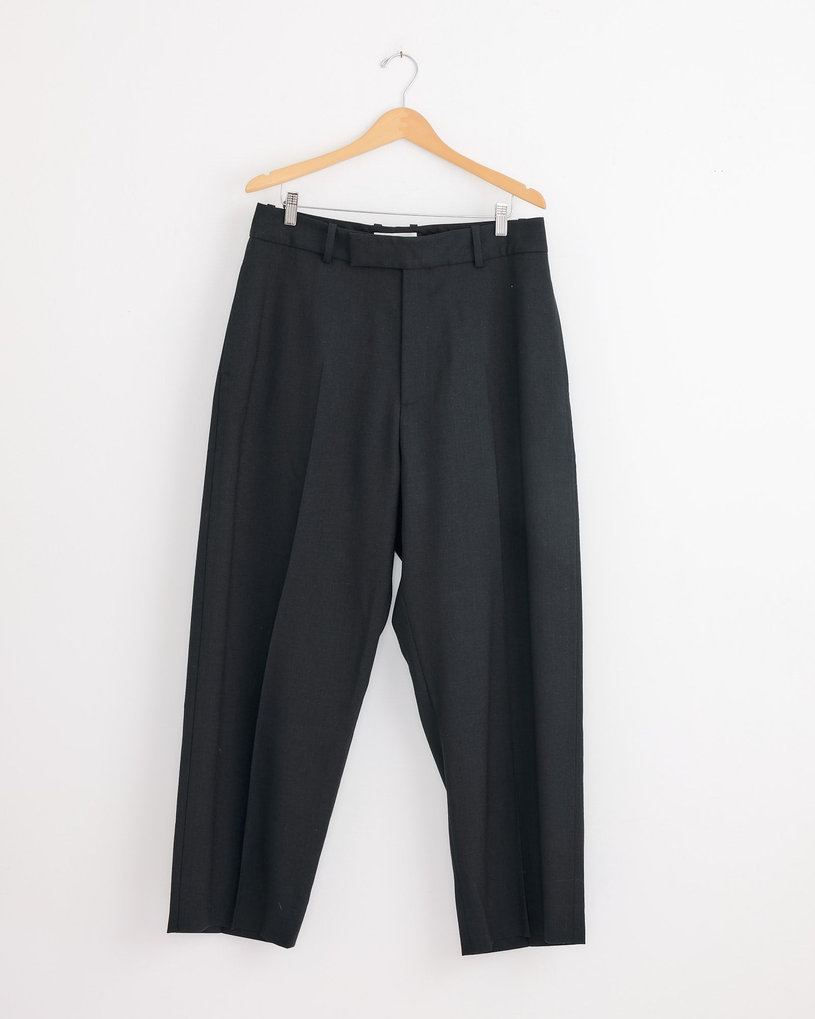 The Studio Nicholson Ester Pant in Darkest Navy, featuring belt loops and a tailored wool design, hangs on a wooden hanger against a white wall.
