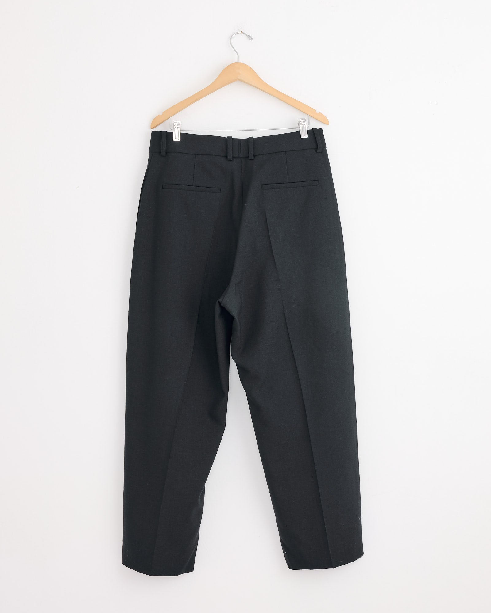 A pair of Studio Nicholson Ester Pants in Darkest Navy—tailored wool trousers in Italian-milled grain de poudre—hang on a wooden hanger against a plain white wall, showing belt loops and two back pockets.