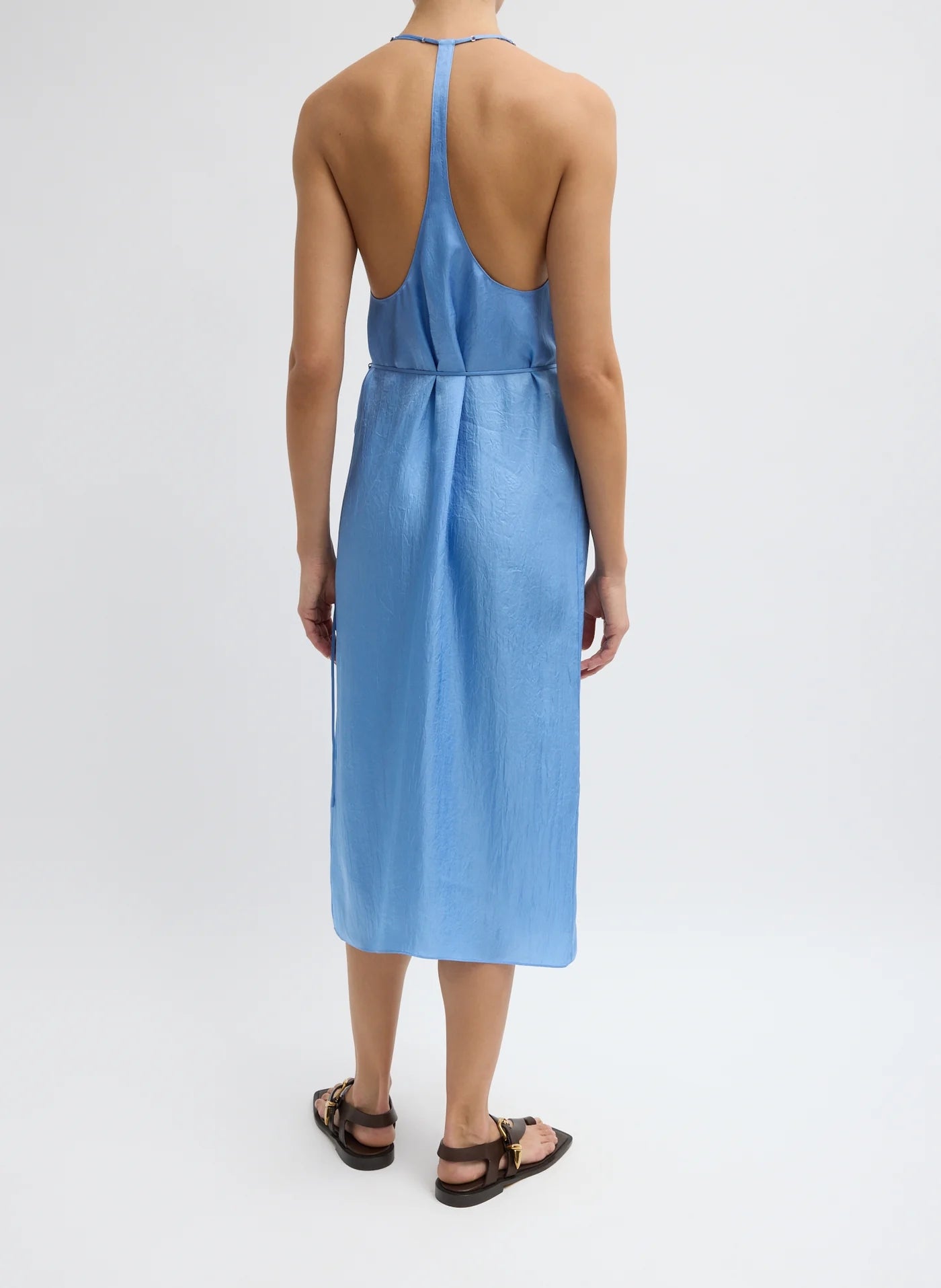 A woman stands facing away in the Tibi Slip Wrap Dress in Baby Blue, a chic sleeveless racerback style, paired with brown sandals against a plain white background—perfect for summer.