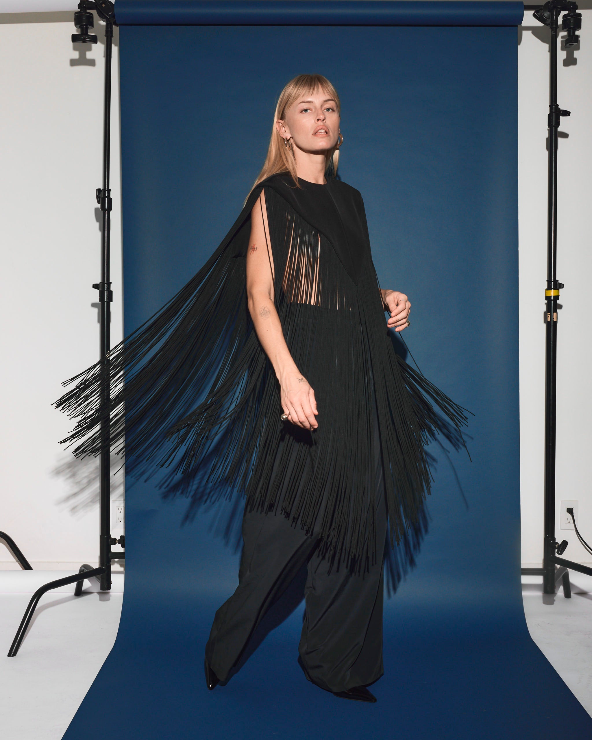 A person poses confidently before a blue backdrop, wearing Tibi's 4-Ply Silk Fringe Cape in Black. Their straight blond hair and dangling earrings complement the cape's long, flowing fringe as they stand with one hand at their waist.