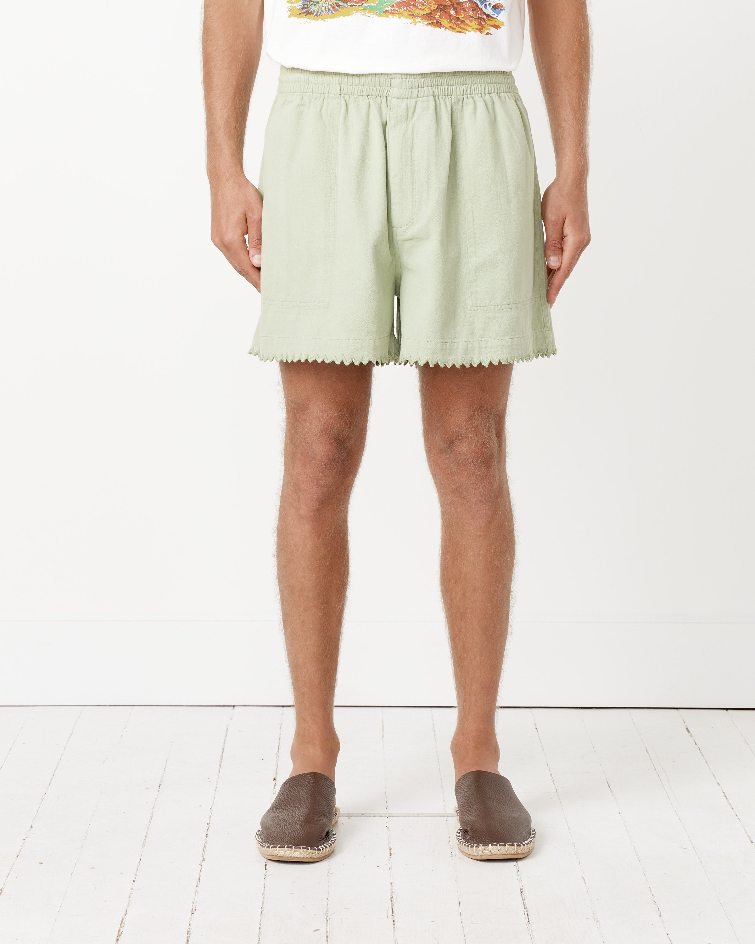 A person stands against a white wall wearing Bode Zig-Zag Hem Shorts, a colorful graphic T-shirt, and brown slip-on shoes. The image is cropped at the shoulders.