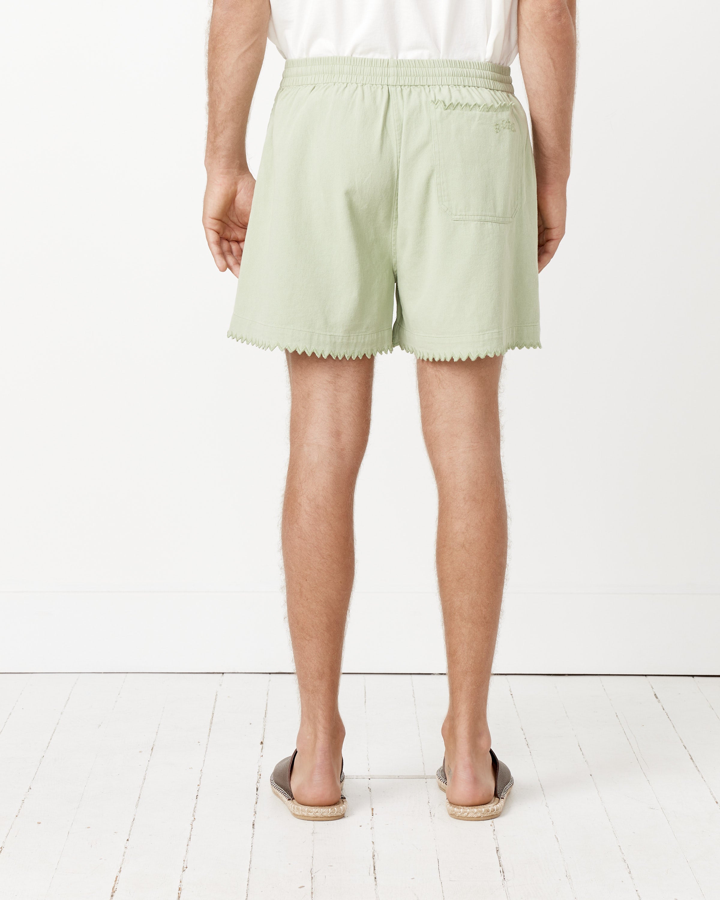 A person faces away, wearing Bode Zig-Zag Hem Shorts in light green with a scalloped hem, paired with a white T-shirt and brown slip-ons on a white wooden floor against a plain white wall.
