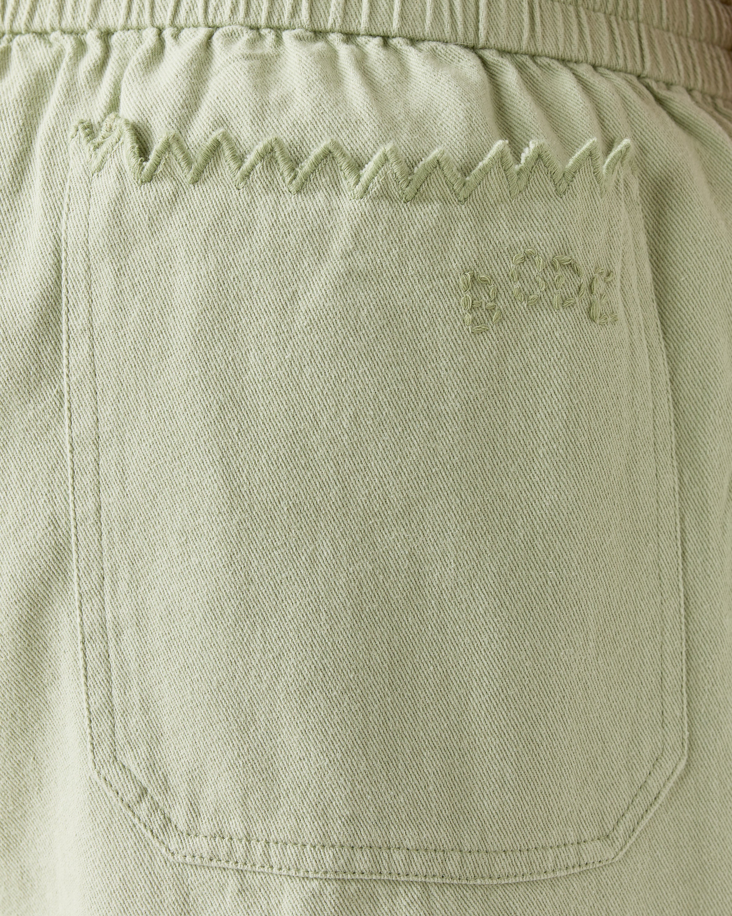 Close-up of Bode's Zig-Zag Hem Shorts shows a light green denim pocket with zigzag stitching along the top edge and faint embroidery on the upper right.