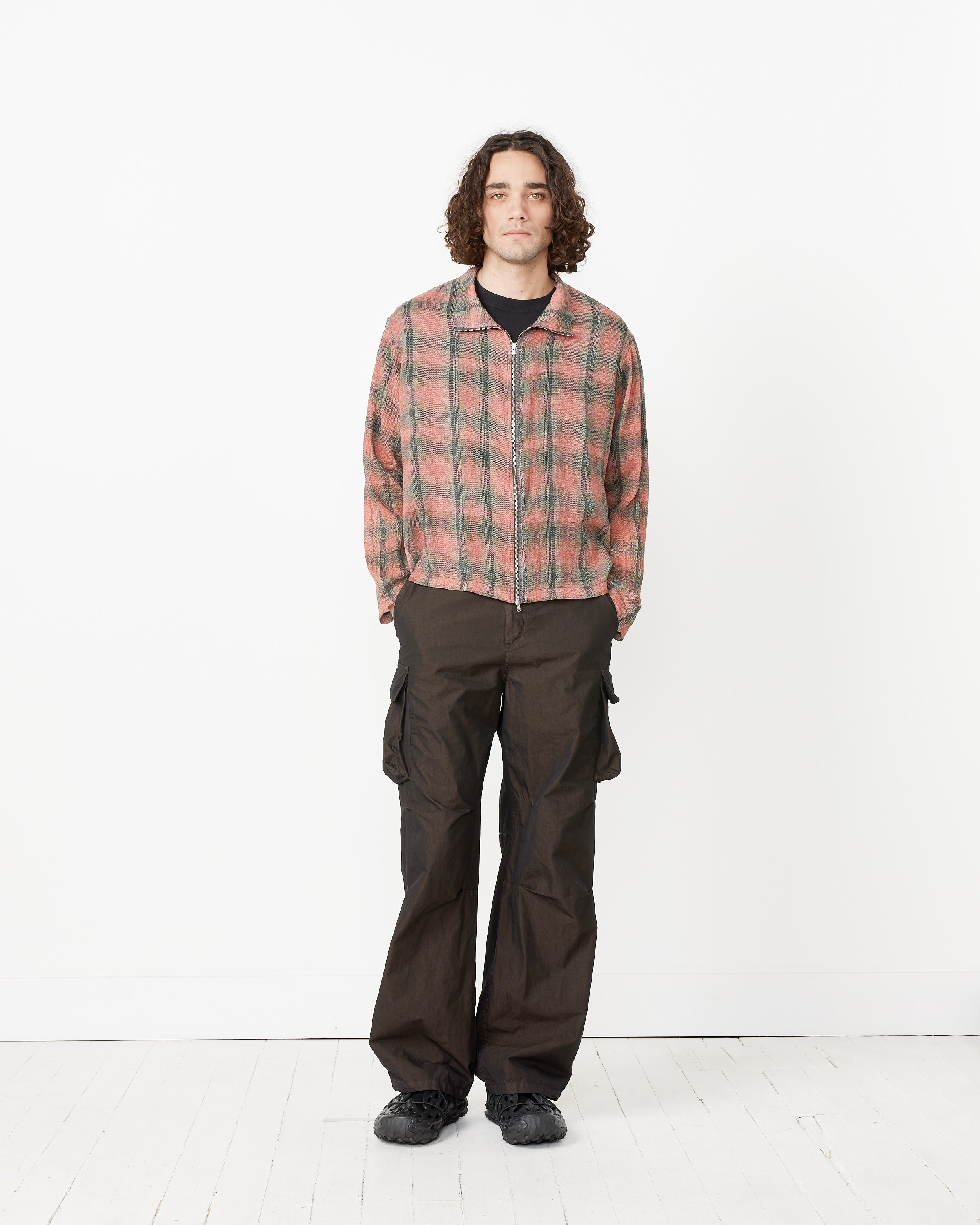 A person with curly hair stands against a white background, wearing the Shrunken Fullzip Polo by Our Legacy over a black t-shirt, paired with dark cargo pants and black shoes.