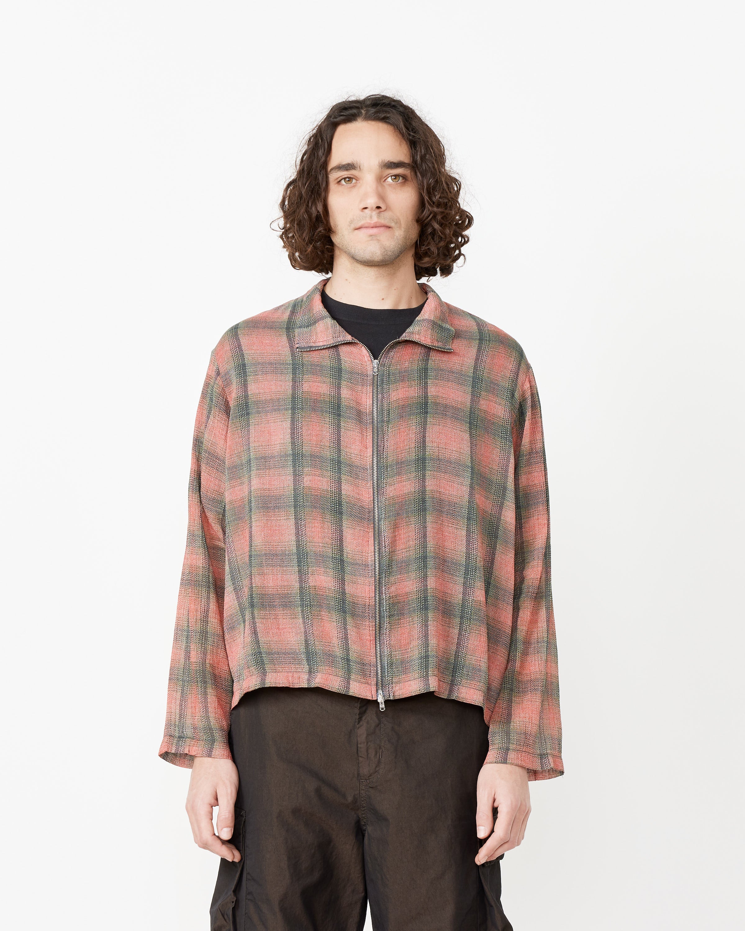 A person with shoulder-length curly hair wears Our Legacy's Shrunken Fullzip Polo in pink and green plaid over a black shirt with loose black pants, standing against a plain white background.