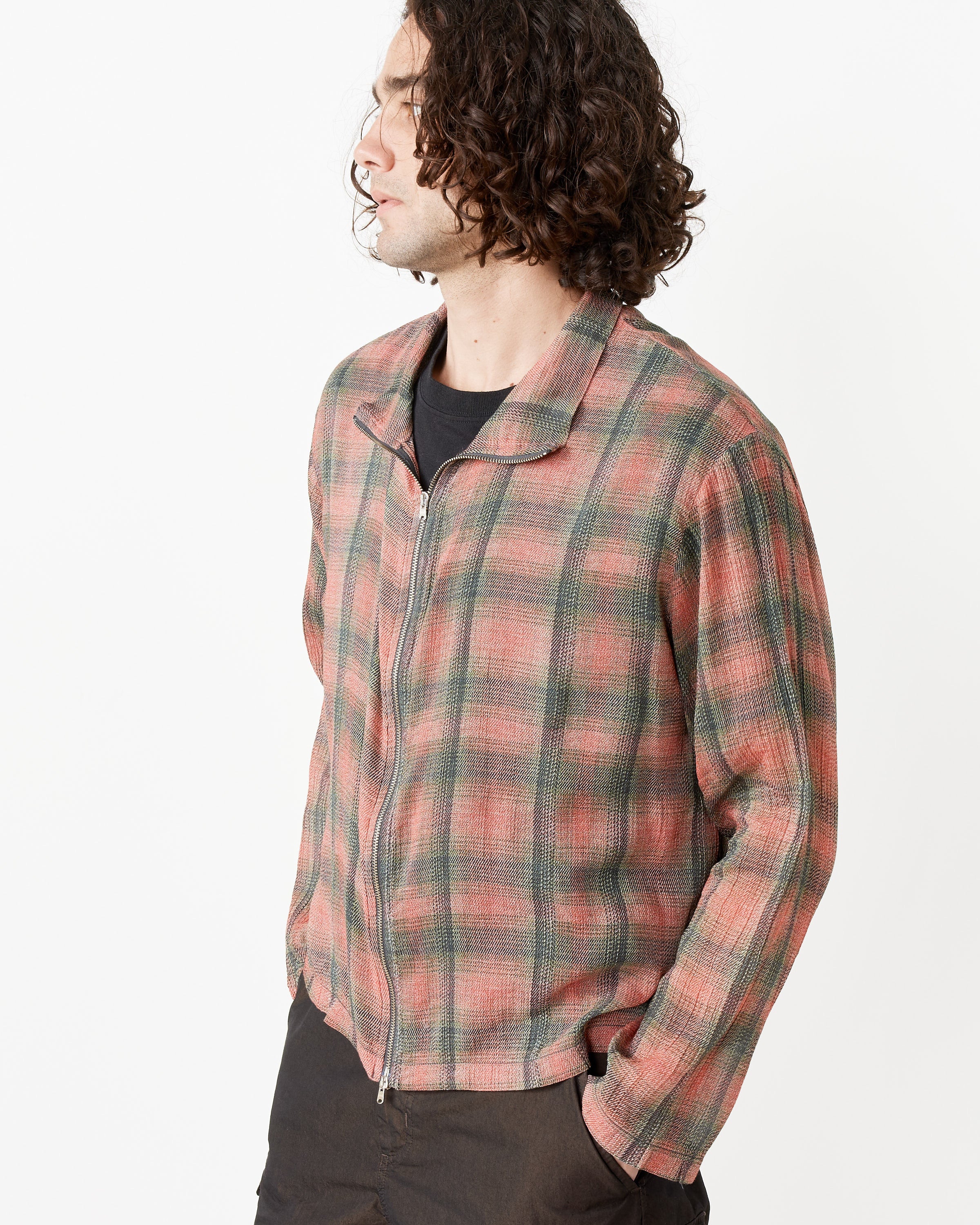 A person with curly, shoulder-length hair models the Our Legacy Shrunken Fullzip Polo in muted red and green plaid over a black shirt and dark pants, standing against a plain white background and looking to the side.