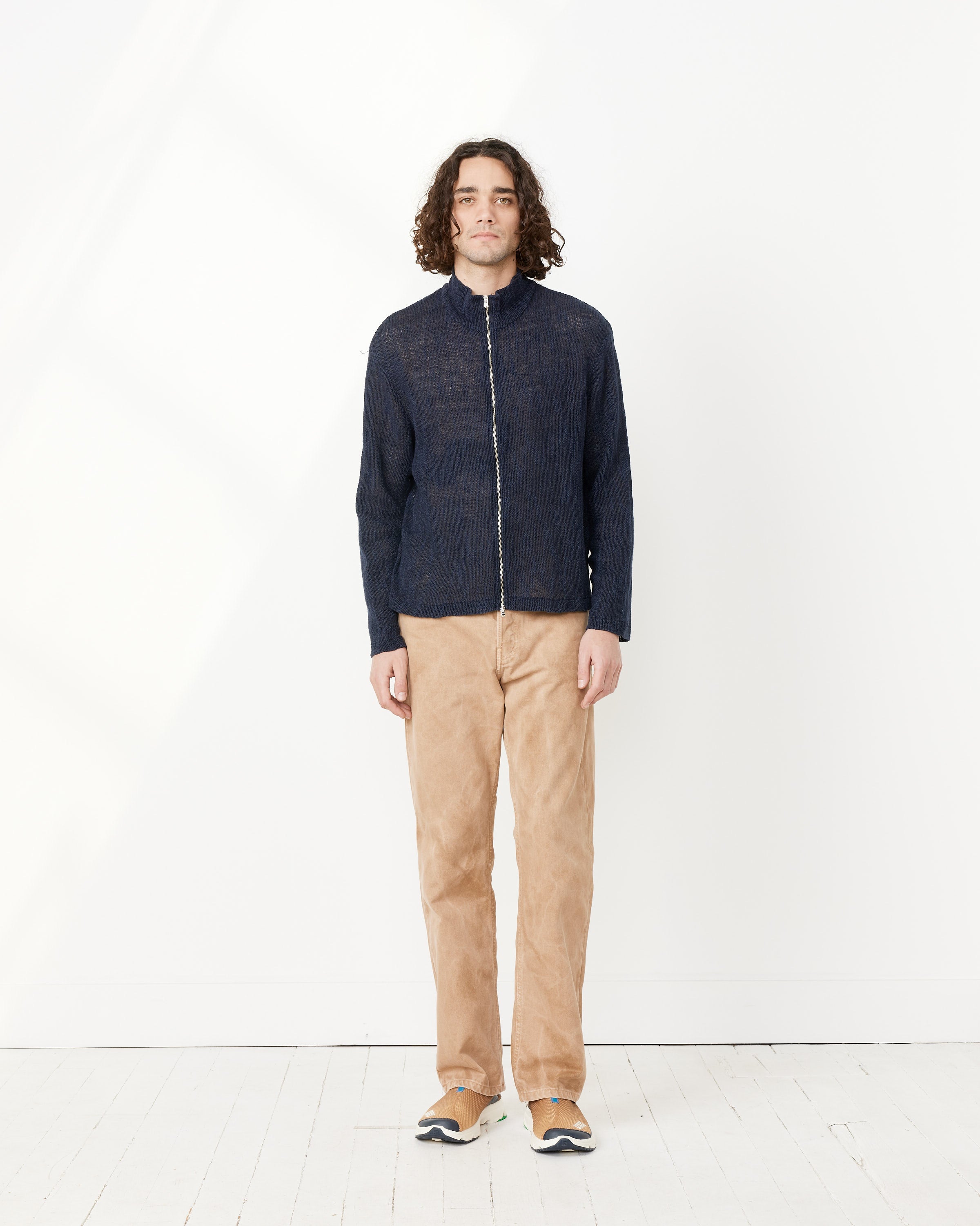 A person with shoulder-length curly hair stands against a plain white wall, wearing the Our Legacy Shrunken Fullzip Polo in navy, paired with beige pants and tan sneakers. The minimal, well-lit background highlights the modern silhouette.
