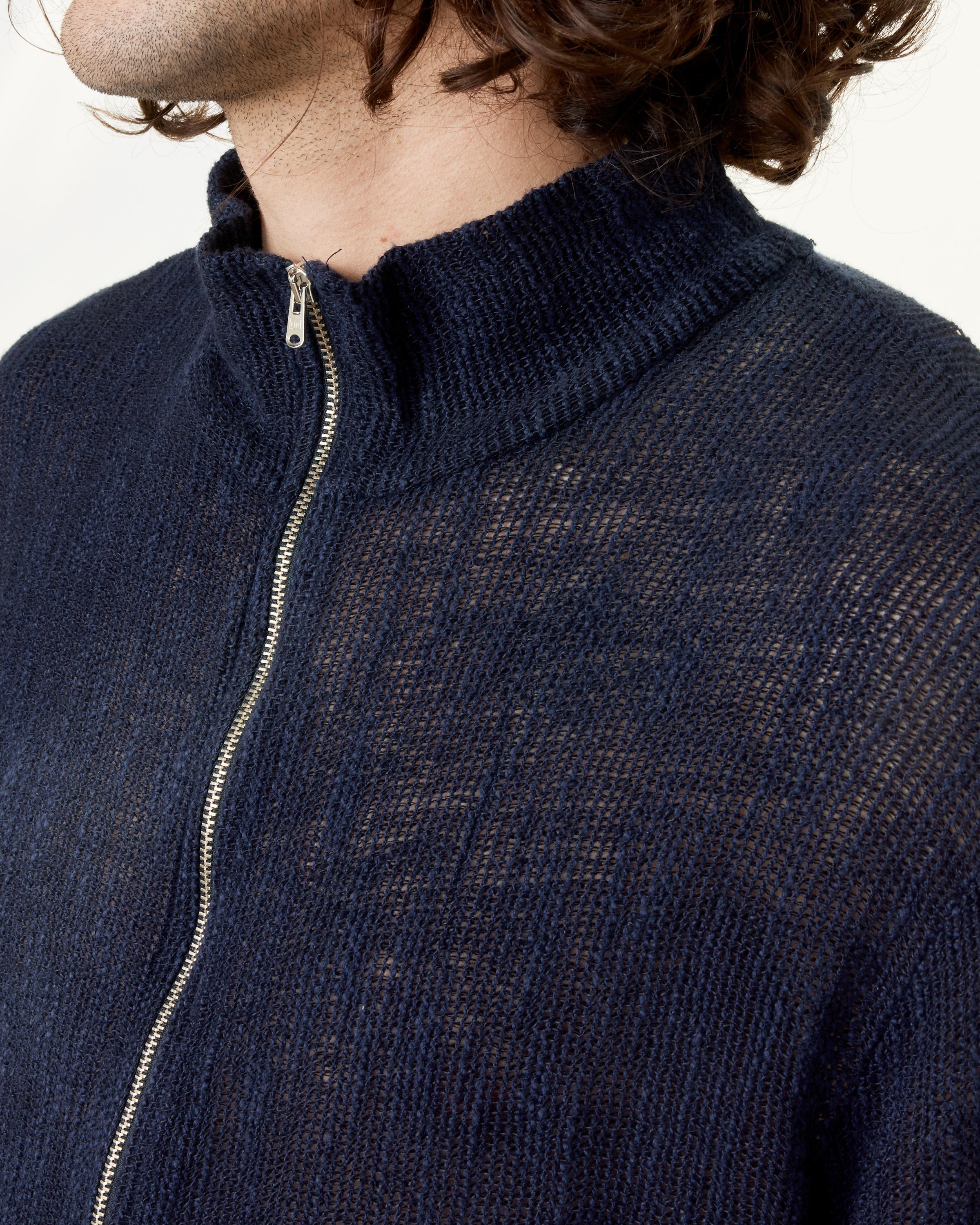 A person with wavy brown hair wears the Our Legacy Shrunken Fullzip Polo, a dark blue, slightly sheer cotton mesh sweater with a high collar and partially closed zipper. The background is plain and light.
