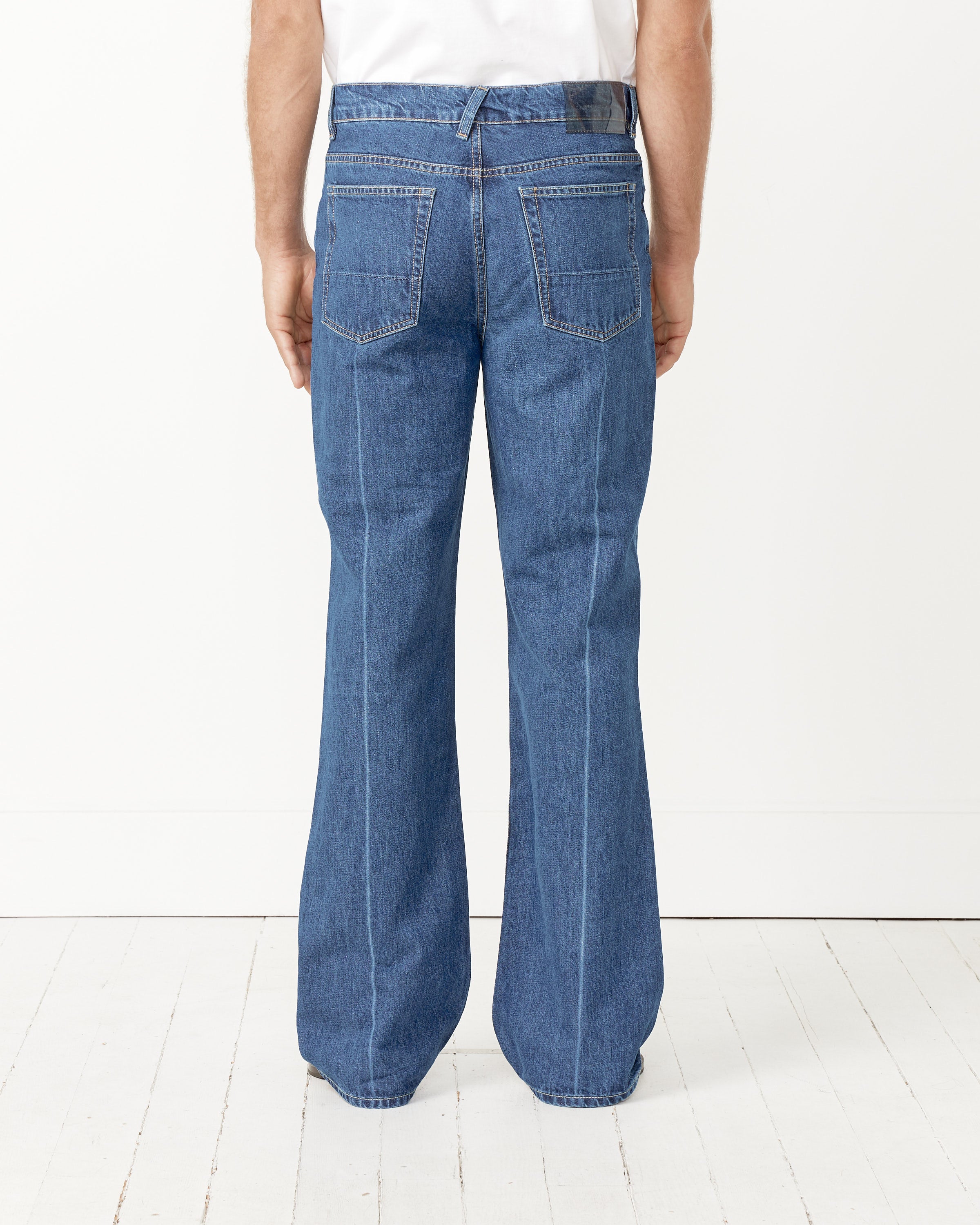 A person stands with their back to a white wall, wearing Our Legacy 70's Cut blue high rise bootcut jeans with large rear pockets, visible seams, and flared legs, paired with a white shirt.