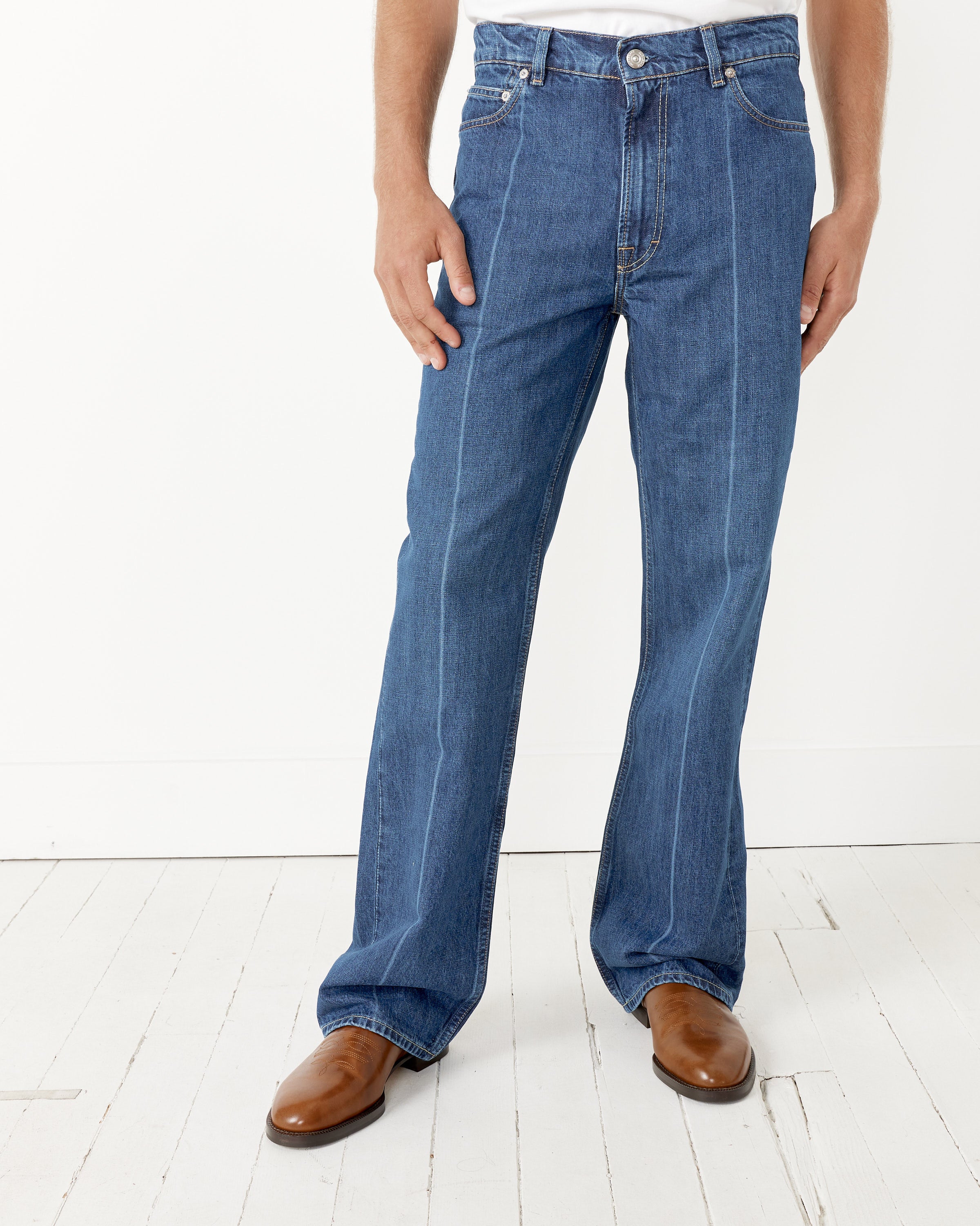 A person wears Our Legacy 70's Cut blue organic cotton denim straight-leg jeans with visible front creases and contrast stitching, paired with brown boots, standing on a white wood floor against a plain white wall.