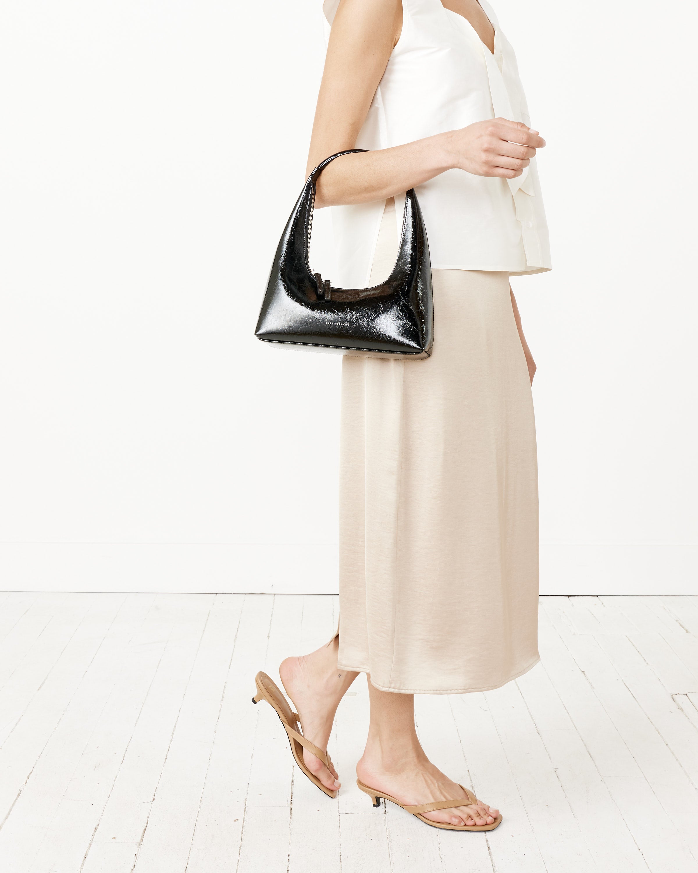 A woman in a white sleeveless top, beige midi skirt, and tan heeled sandals carries the Marge Sherwood Hobo Shoulder Bag - Black Crinkle while standing against a white background.