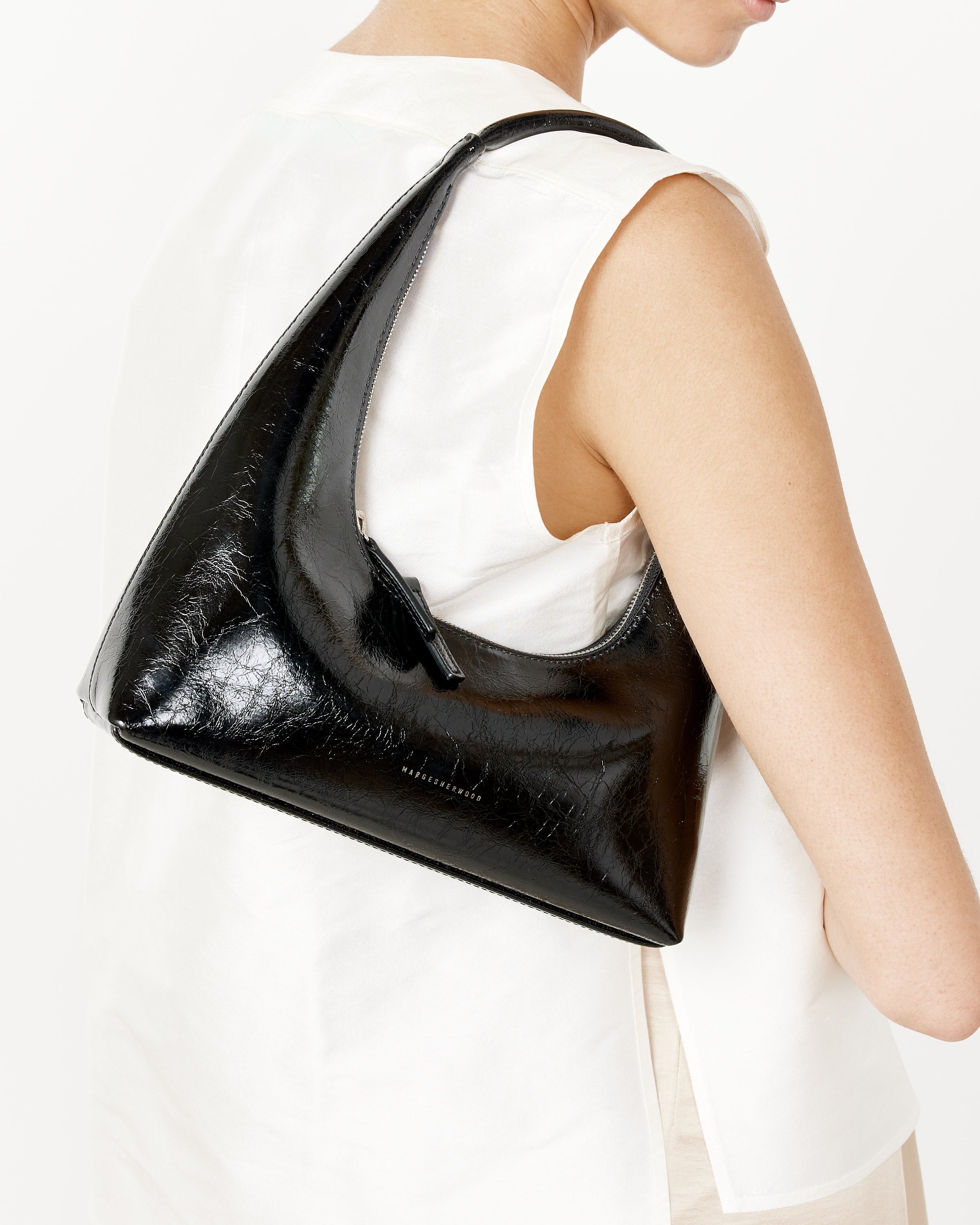 A person in a sleeveless white top is seen from the side carrying the Marge Sherwood Hobo Shoulder Bag - Black Crinkle with a curved silhouette and zipper closure, its glossy Italian cowhide standing out against a plain white background.